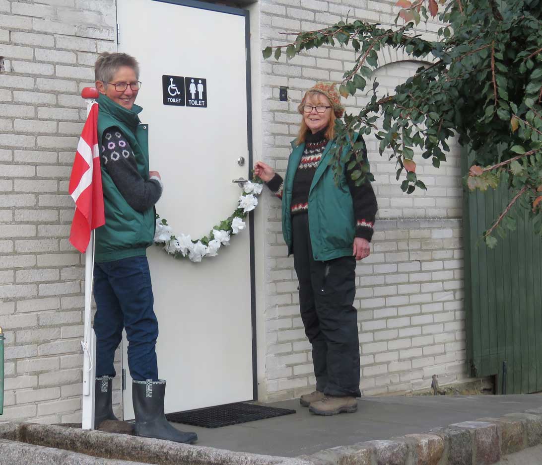 Bente Wulff og Gunvor Rosell hænger efeuranken på plads foran indgangen til toilettet, så den er klar til at blive klippet af Bo Nissen, formand for Trafik- Teknik- og Miljøudvalget i Langeland Kommune, 
